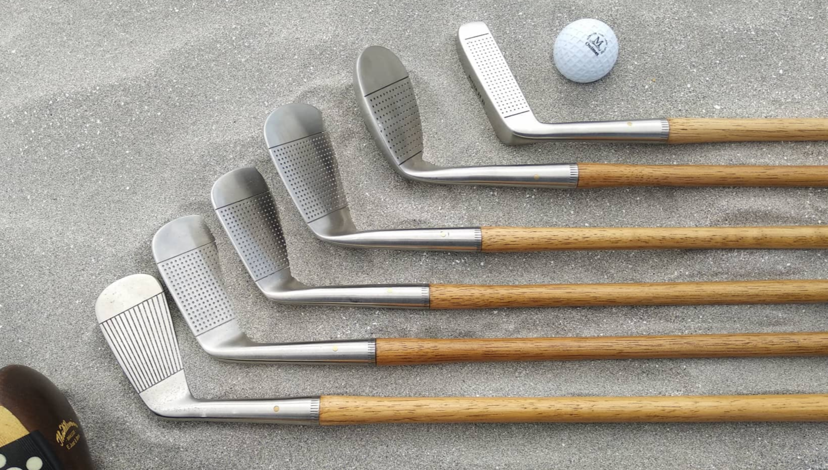 Hickory golf clubs laid together on sand with replica ball.